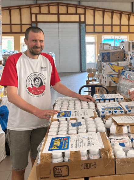 Tara is standing next to loads of supplies in a tin building.