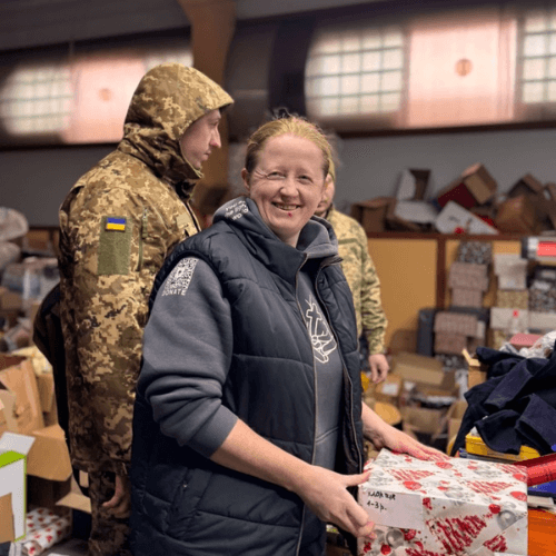 Valentyna smiles while holding a wrapped gift in a humanitarian aid warehouse in Ukraine, helping prepare Christmas presents and relief supplies for children and families affected by the war.