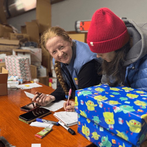 Valentyna works with another volunteer to record and organize Christmas gifts for children in Ukraine, preparing humanitarian aid packages at a busy warehouse filled with donations.