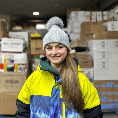 A woman stands in a humanitarian aid warehouse in Ukraine with boxes of donated supplies behind her, volunteering to help organize and distribute relief items for families affected by the war.